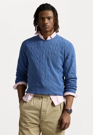 Pull en maille bleu superposé sur une chemise à col rose clair, associé à un pantalon beige plissé. Montre-bracelet notable visible.