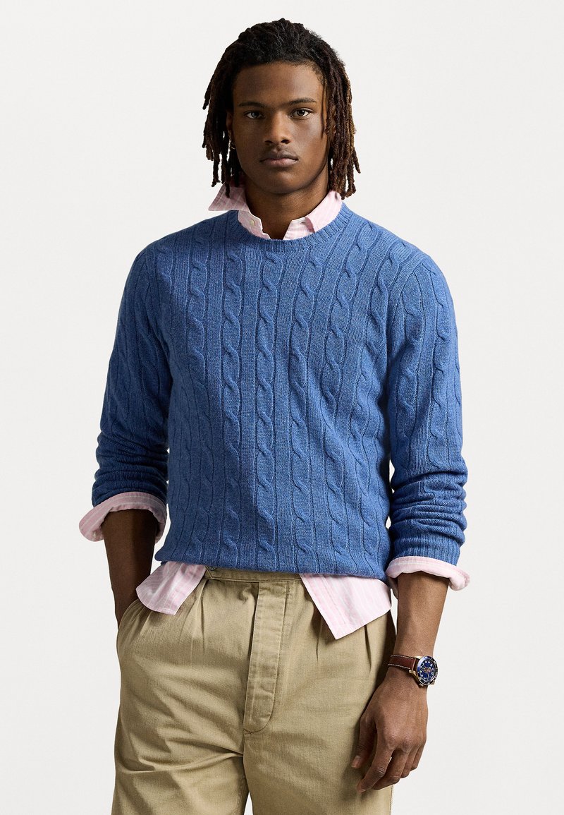 Μπλε πουλόβερ με πλέξη καλωδίου, φορεμένο πάνω από ένα ανοιχτό ροζ πουκάμισο με γιακά, συνδυασμένο με μπέζ πλισέ παντελόνι. Ορατό ρολόι χειρός.