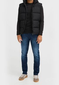 Schwarze gepolsterte Weste mit Kapuze über einem schwarzen Pullover, kombiniert mit blauen Jeans und grauen Sneakers mit weißen Akzenten und Schnürsenkeln.