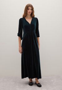 Robe en velours verte foncée, longue, avec un décolleté en V, des manches bouffantes et une taille élastique. Associée à des chaussures plates noires avec des accents décoratifs.