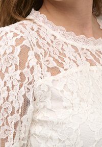 Top bianco in pizzo con motivi floreali, scollatura a scalloped, tessuto trasparente e design a strati. Vista ravvicinata che mette in evidenza i dettagli intricati.