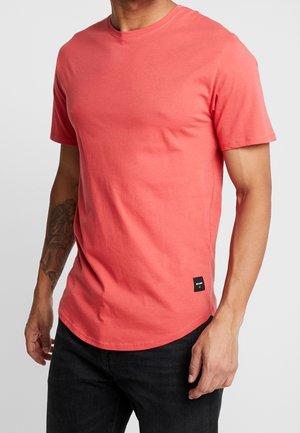 Mand iført simpel koralfarvet T-shirt med korte ærmer og sorte bukser, med en synlig tatovering på venstre underarm mod en ensfarvet baggrund.