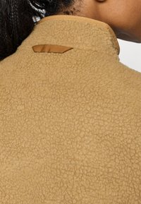Gros plan d'une personne portant une veste en polaire texturée couleur beige avec une boucle en tissu marron au niveau du col arrière.