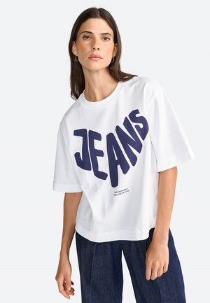 T-shirt blanc surdimensionné avec "JEANS" en grandes lettres bleu marine sur le devant ; encolure ronde et manches courtes, fabriqué en tissu de coton.