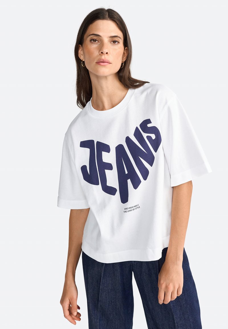 T-shirt blanc surdimensionné avec "JEANS" en grandes lettres bleu marine sur le devant ; encolure ronde et manches courtes, fabriqué en tissu de coton.