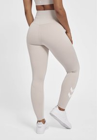 Beige actieve leggings met een hoge taille, een soepele textuur en witte chevronaccenten op het onderbeen, in combinatie met witte sportschoenen.