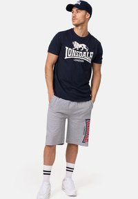 Tricou bleumarin cu logo alb "Lonsdale London", combinat cu pantaloni scurți gri cu accente roșii și logo pe lateral. Purtați cu adidași albi și șosete în dungi.