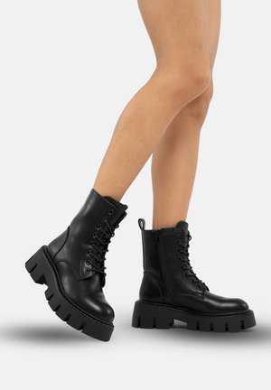 Des bottes chelsea en cuir noir présentent un laçage à l'avant et une fermeture éclair sur le côté, avec une semelle en caoutchouc épaisse et une finition texturée.