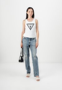 Un top alb ajustat fără mâneci, cu un logo triunghiular negru, blugi decolorati de culoare albastru deschis, sandale crem cu talpă plată și o geantă de mână neagră.