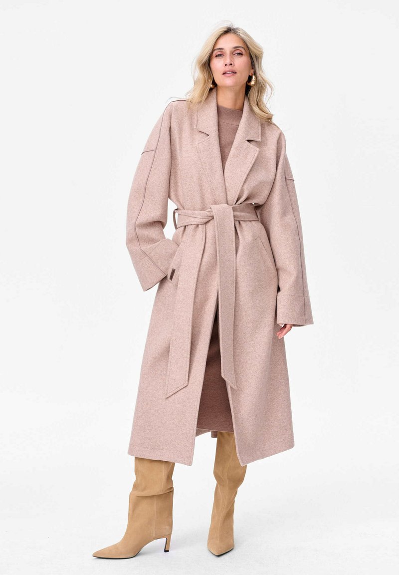 Długi płaszcz z paskiem w miękkim różowym materiale; posiada szerokie rękawy, kołnierz z klapami oraz boczne kieszenie. Zestawiony z beżowymi botkami do kolan.