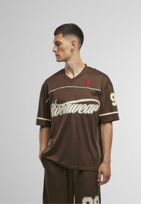 Barna rövid ujjú póló lélegző anyagból, fehér és piros díszítéssel, V-nyakkal, és nagy "Rootwear" logóval, 98-as számmal.