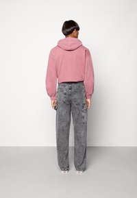 Sudadera con capucha rosa combinada con pantalones cargo de mezclilla negra, holgados y deslavados, que cuentan con múltiples bolsillos. Fondo liso.