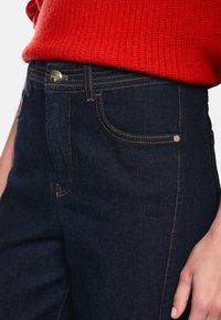 Dunkelblaue Jeans aus Denim mit orangefarbener Naht. Verfügt über einen Metallknopf und Taschen auf der Vorderseite. Oben ist ein roter Strickpullover zu sehen.