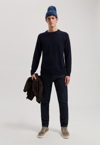 Marineblauwe gebreide trui met geribde manchetten, donkere denim jeans, beige en grijze sneakers, en een blauwe beanie. Bruine jassen vastgehouden in de linkerhand.