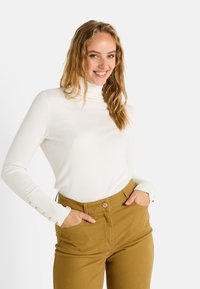 Jersey de cuello alto blanco hecho de un material suave, con mangas largas y detalles de botones. Combinado con pantalones de alta cintura en color caqui.