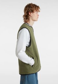 Gilet in pile verde con scollo rotondo, rifiniture nere, tasche laterali e vestibilità comoda, indossato sopra una camicia bianca a maniche lunghe.