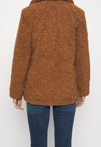 Brun fleecejacka med en texturerad, fluffig yta och huva. Har långa ärmar och en avslappnad passform som sträcker sig till höften.