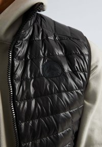 Gilet en duvet noir avec sections cousues, texture brillante, fermeture éclair et un patch logo circulaire sur la zone de la poitrine gauche.