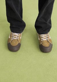 Brune semskede sneakers med hvite detaljer, har tykke snørebånd og gummisåler. Kombinert med mørk denimbukser, satt mot en grønn bakgrunn.
