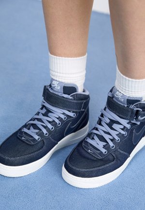 Stopy w granatowych wysokich sneakersach Nike Air Force 1 z jasnoniebieskimi sznurówkami i białych skarpetkach na jasnoniebieskiej powierzchni.