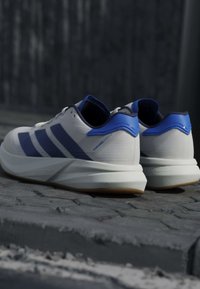 Chaussures de course blanches avec des rayures bleues et des accents au talon étiquetées "Duramo Speed 2" reposant sur une surface extérieure pavée.
