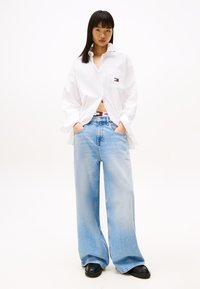 Chemise blanche oversize à boutons avec une poche poitrine, associée à un jean bleu clair à jambe large et des chaussures à plateforme noires.