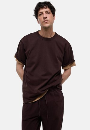 Homme aux cheveux bouclés et foncés portant des t-shirts marron superposés et un pantalon marron à cordon assorti, debout les mains derrière le dos.