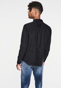 Svart långärmad skjorta, texturerad tyg, knapplånga ärmar, böjd fåll, kombinerad med blekta blå jeans, står mot en vit bakgrund.