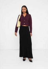 Avskuren vinröd cardigan med knappar, tillsammans med en svart maxikjol. Modellen bär en ljusgul väska. Svarta klackskor fulländar outfiten.