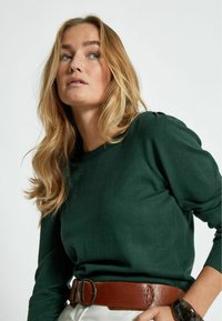 T-shirt en maille vert à manches longues avec encolure ronde et manches bouffantes, associé à une ceinture marron avec une grande boucle ronde.