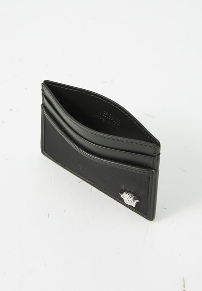 Black leather cardholder with smooth texture, two slanted pockets, and a small silver emblem accent on the front. Compact and minimal design.