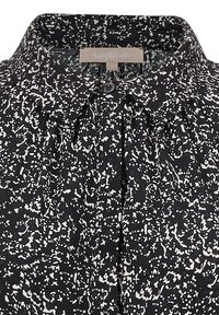 Blusa nera con un motivo astratto bianco, caratterizzata da un colletto plissettato e una chiusura con bottoni. Il materiale è liscio e leggero.