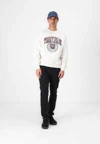 Witte sweatshirt met "TOMMY JEANS" grafiek, zwarte jeans en zwarte sneakers. Blauwe pet maakt de outfit compleet. Model staat op een effen achtergrond.