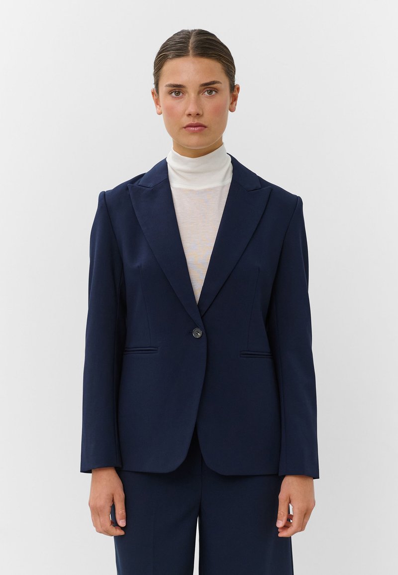 Γυναίκα με σκουρόχρωμα μαλλιά που είναι πιασμένα πίσω, φοράει ένα σκούρο μπλε σακάκι πάνω από ένα λευκό ζιβάγκο, με φόντο ένα απλό ανοιχτό χρώμα.