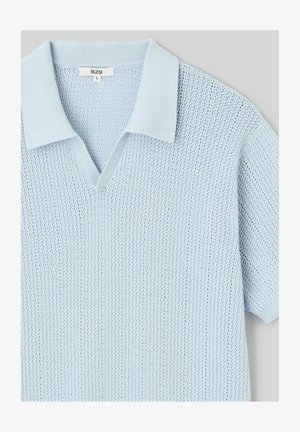 Polo en maille bleu clair à manches courtes avec col en V et col et poignets côtelés, étiquette de taille visible à l'intérieur du col.