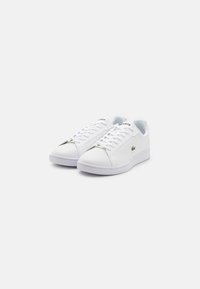 Een paar witte Lacoste lage sneakers met veters en een groen krokodillenlogo aan de zijkant, geplaatst op een effen witte achtergrond.