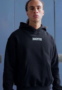 Čierna mikina s kapucňou a predným vreckom; obsahuje logo "JUVENTUS" v bielej farbe napísané cez hruď. Mäkká textúra, pohodlný strih.