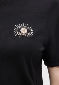 Μαύρο κοντομάνικο πουκάμισο με λευκό κεντητό σχέδιο ματιού στο στήθος, που περιλαμβάνει βλεφαρίδες και κυκλικά στοιχεία.