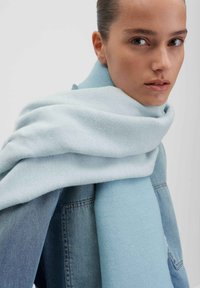 Sciarpa blu chiaro realizzata in tessuto morbido, elegantemente drappeggiata attorno al collo, con una consistenza liscia e una leggera variazione di colore, abbinata a una giacca di denim.