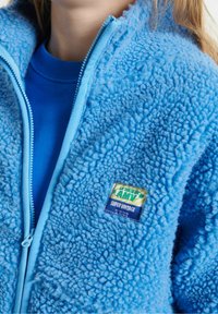 Chaqueta de piel sintética azul con cierre de cremallera y una textura suave. Presenta una etiqueta con un diseño vintage. Debajo, se puede ver una camisa azul sólida.