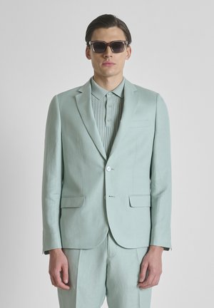 Man die een lichtgroen pak draagt en een bijpassend geribd poloshirt met grote donkere zonnebril, staand tegen een effen grijze achtergrond.