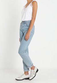 Top bianco senza maniche con pizzo, jeans leggeri a vita alta di colore blu chiaro e scarpe bianche con borchie e fibbie. Design semplice, look casual.
