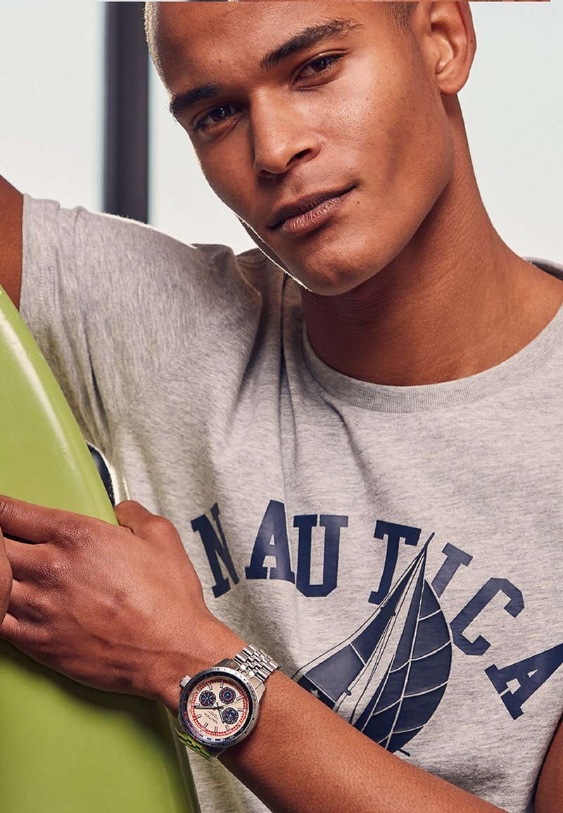 Silverfärgad armbandsklocka med vit urtavla och blå accenter, som bärs på en solbrun arm. Grå t-shirt med blå text och segelbåtsgrafik. Grön surfbräda i bakgrunden.