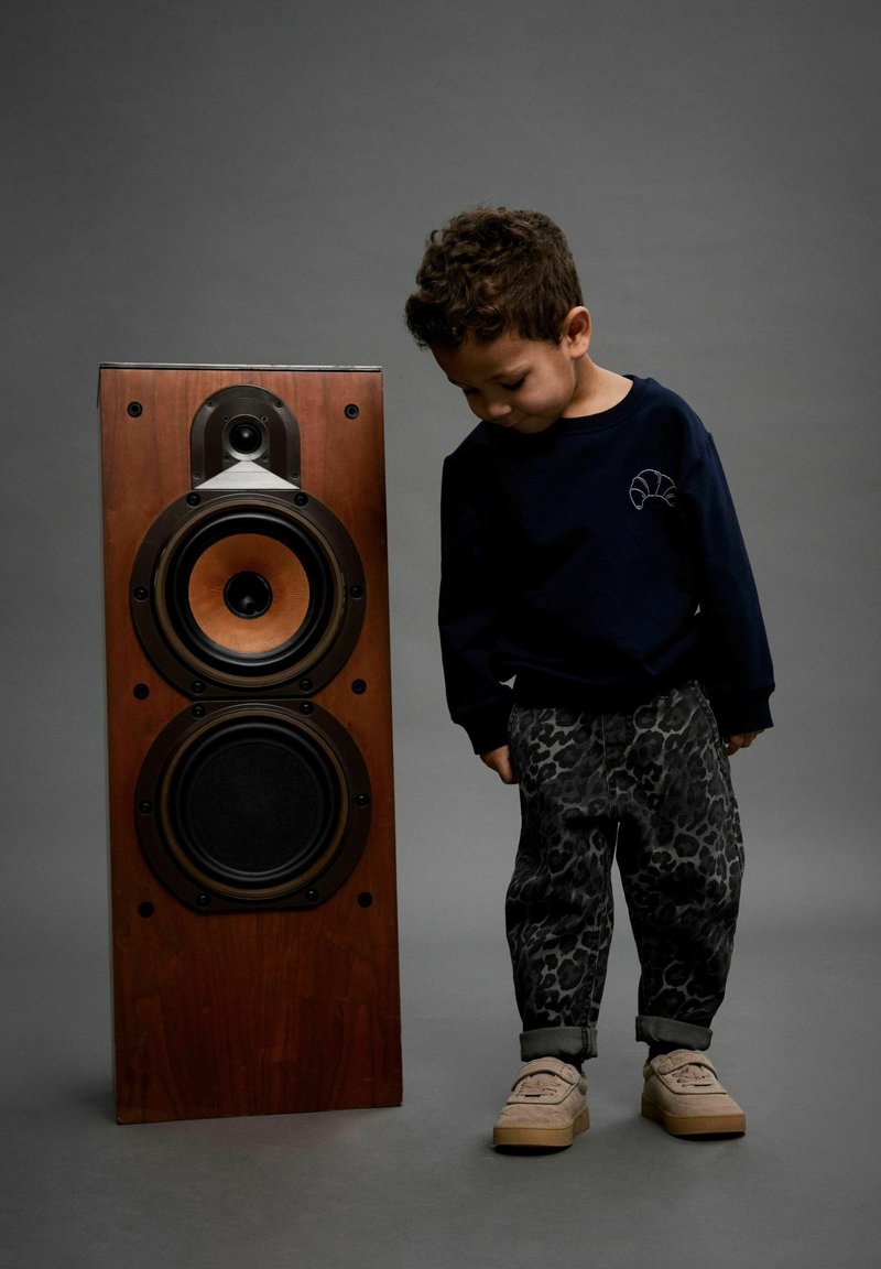 Haut-parleur en bois avec des haut-parleurs noirs et cuivrés; un enfant en sweatshirt sombre et pantalons imprimés léopard, portant des chaussures claires, se tient à côté.