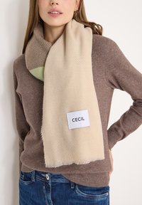 Beige rektangulært tørklæde med fiskebensmønster, der har en grøn sektion og et hvidt mærke med "CECIL" over en brun sweater.