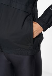 Mano insertada en el bolsillo lateral de una chaqueta ligera negra, llevada sobre leggings negros con un puño de manga elástica visible.