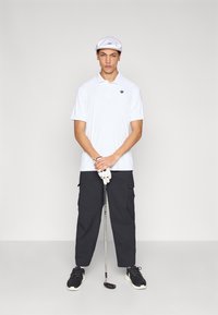 Polo blanc avec un logo, pantalon cargo noir et casquette. Porte un gant de golf et tient un club de golf. Baskets noires.