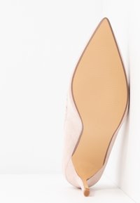 Beige suede pointed-toe stiletto heel shoe positioned sole-up against white corner background.