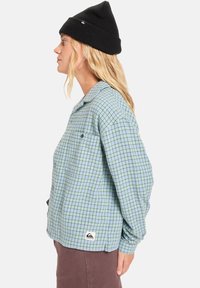 Lichtblauw geruite flanellen shirt met een kraag, zijzak en een logopatch. Draagt een zwarte beanie en toont lange mouwen en een relaxte pasvorm.