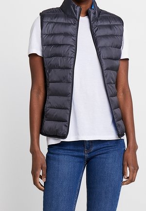 Personne portant une veste matelassée noire sans manches sur une chemise blanche à manches courtes et un jean bleu, debout, les mains détendues le long du corps.